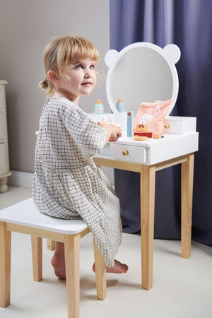 A young girl sits at a white wooden dressing table with a round mirror, playing with toys and beauty products.