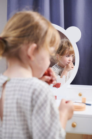 A young girl in a checkered outfit gazes into a round mirror on a white dressing table, applying lip color.