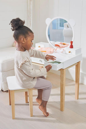 A young girl sits at a white wooden dressing table with a round mirror, pulling out a drawer.