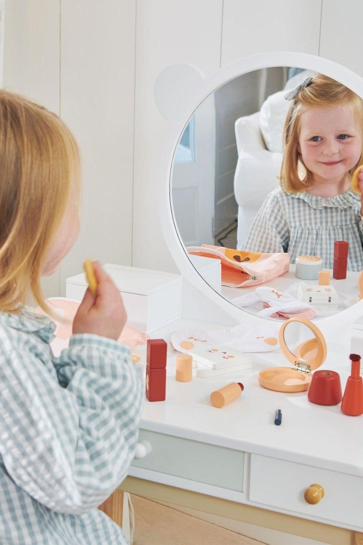 A young girl in a checked dress smiles at her reflection in a round mirror on a white wooden dressing.