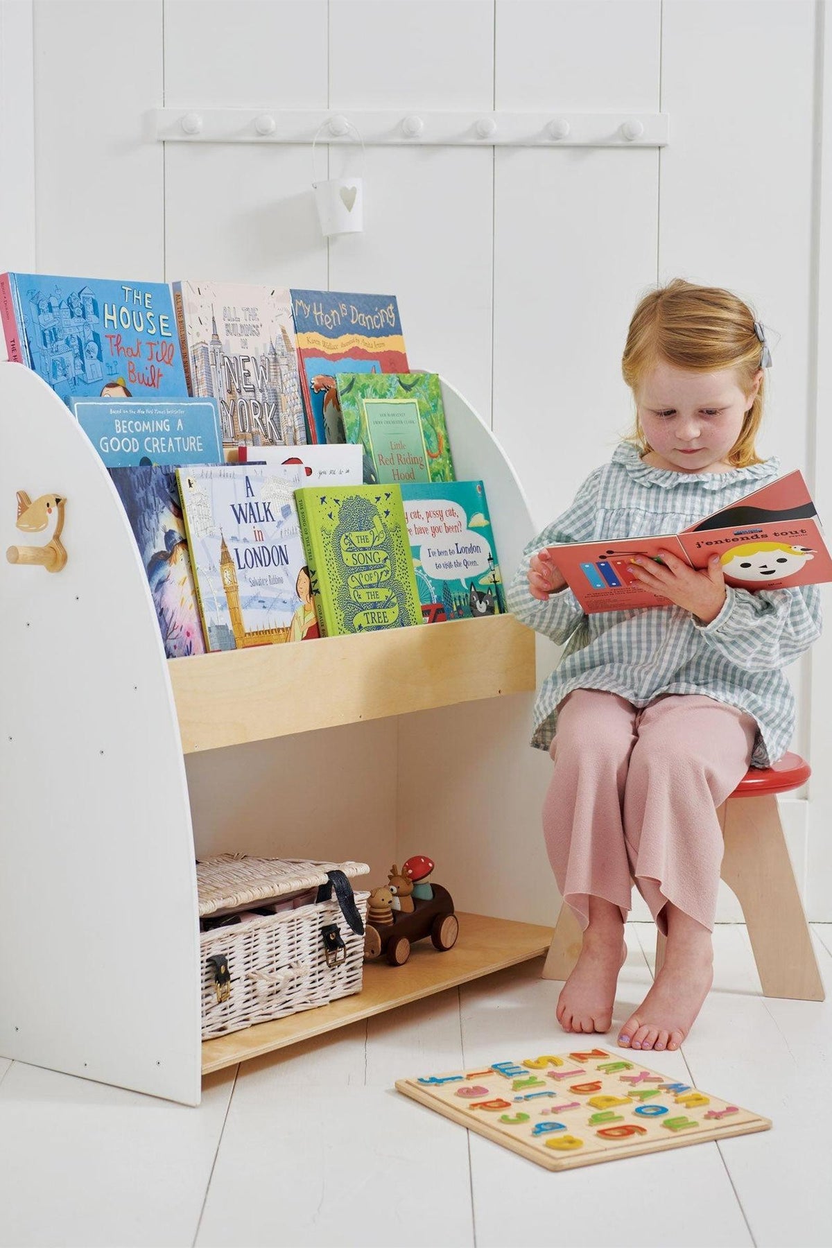 A young girl sits on a red stool, reading a book in front of a white and wood bookshelf filled.