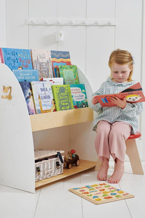 A young girl sits on a red stool, reading a book in front of a white and wood bookshelf filled.