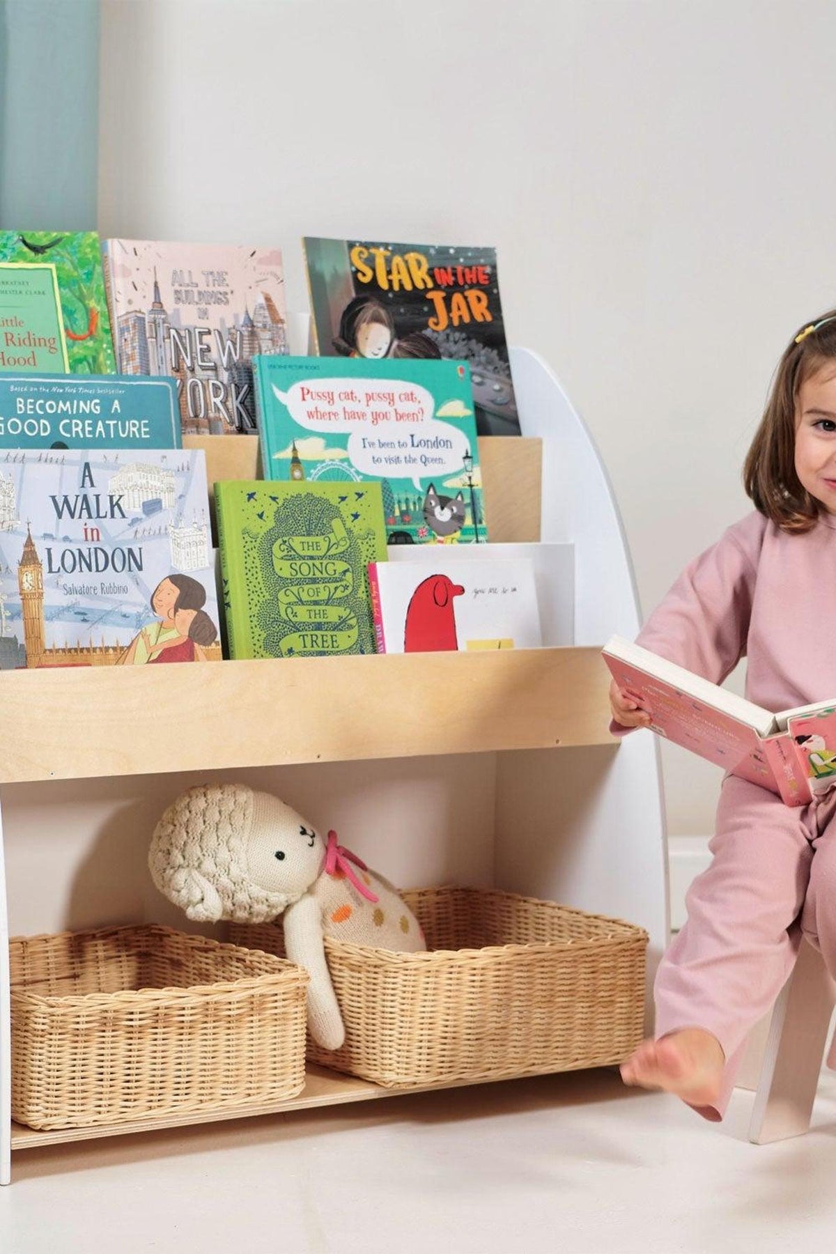 A young girl sits beside a wooden bookshelf filled with colorful picture books and woven storage baskets below.