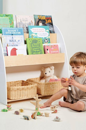 A toddler sits on the floor, playing with colorful wooden toys near a bookshelf filled with childrens books and woven.