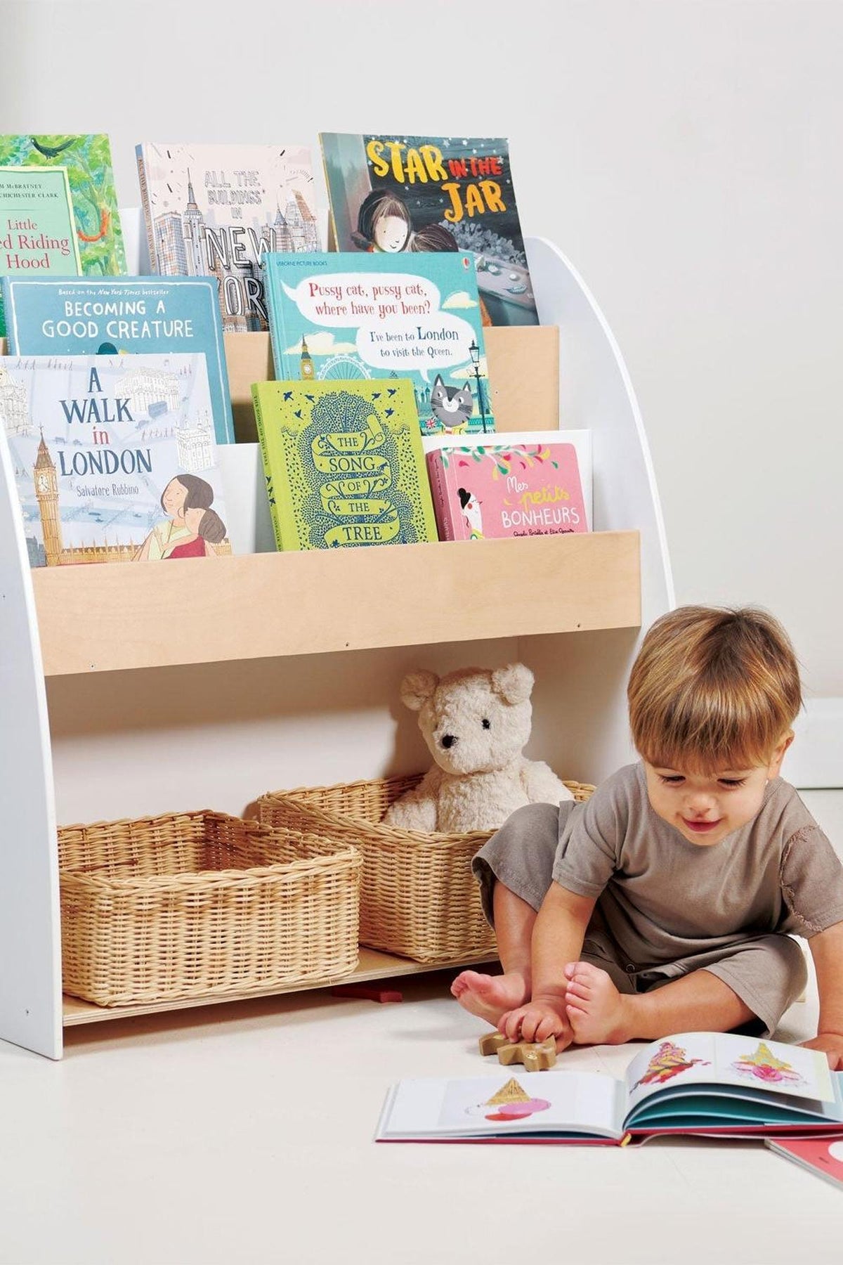 A young child sits on the floor, reading a colorful picture book beside a wooden bookshelf filled with various childrens.