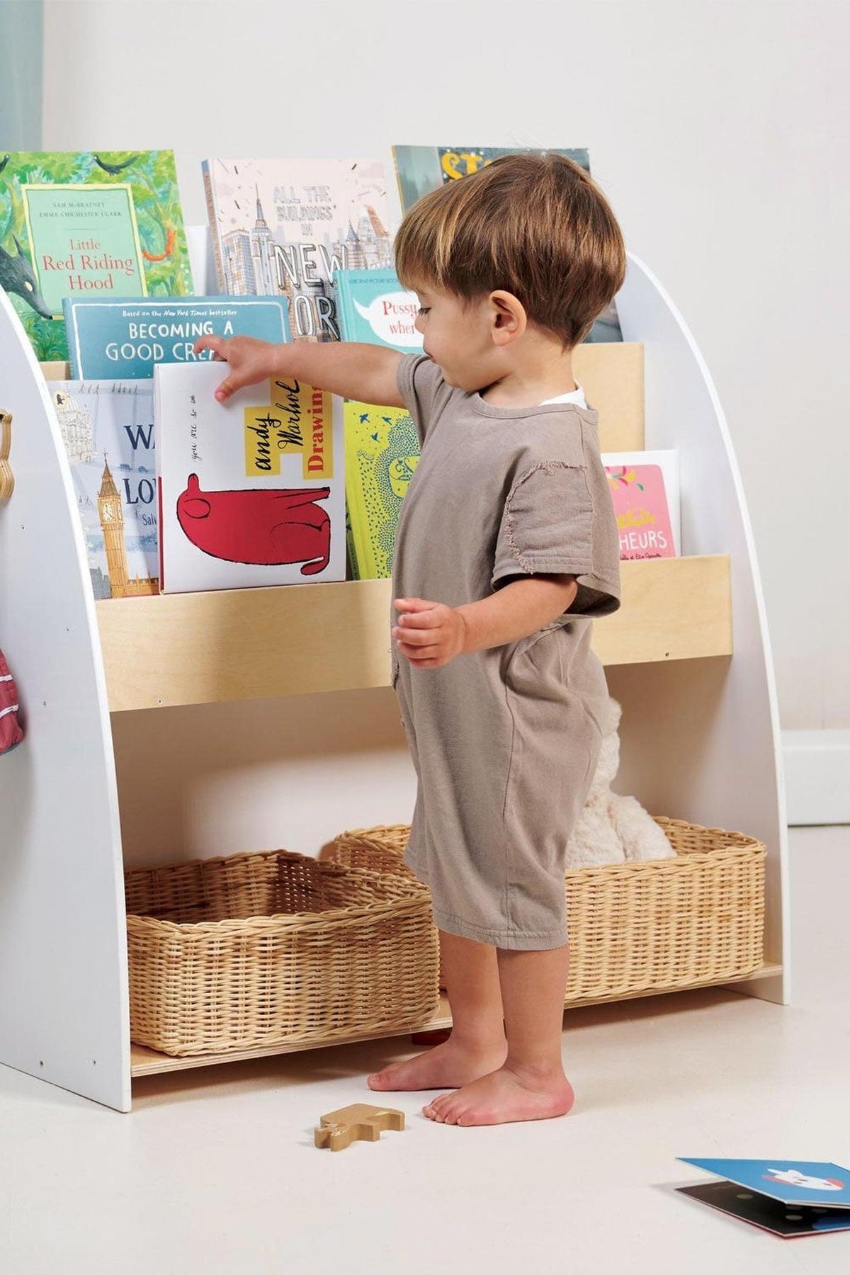 A toddler in a beige outfit reaches for colorful picture books on a white and natural wooden bookshelf, with woven.