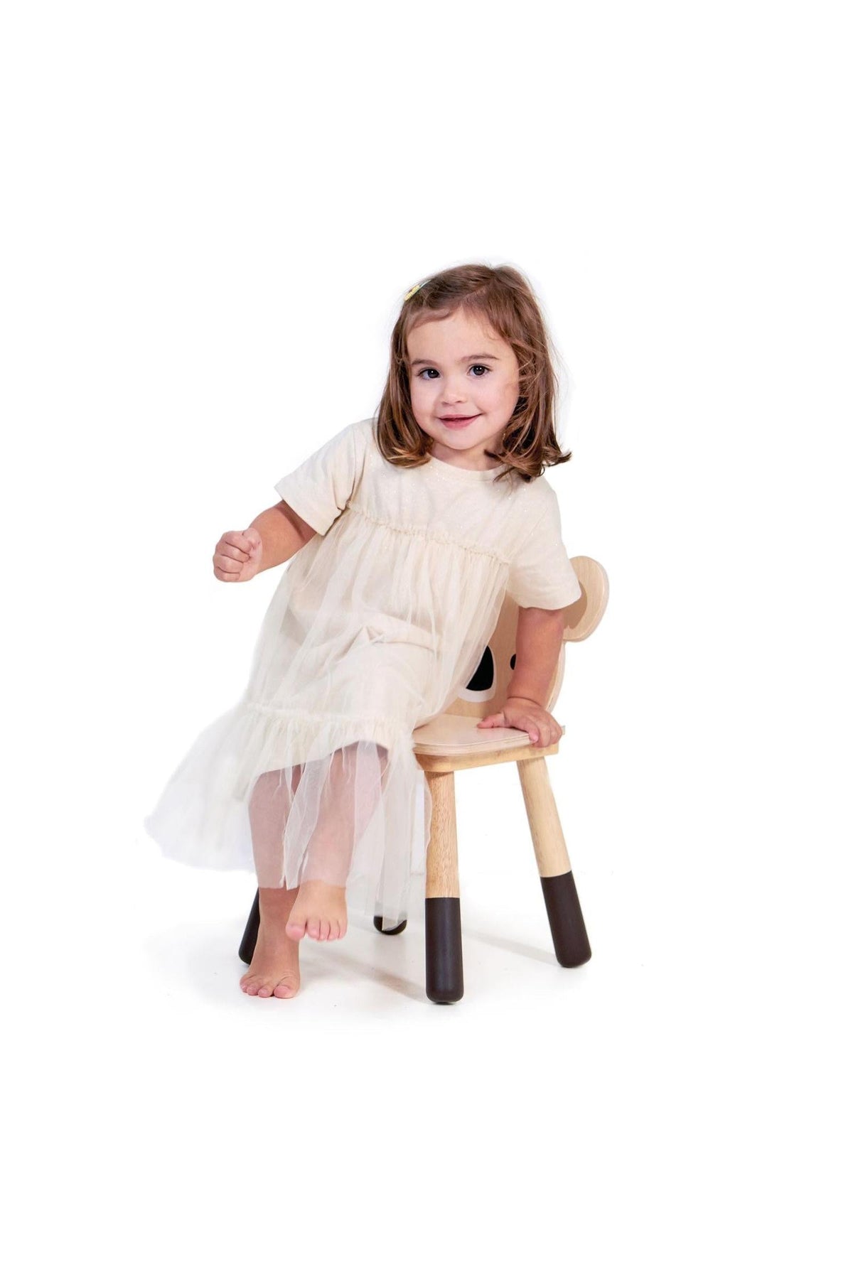 A young girl sits on a wooden chair, wearing a light dress, smiling while playfully posing.