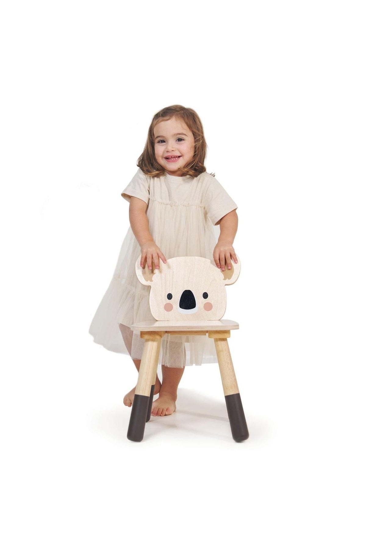 A toddler in a light dress stands beside a wooden koala chair, featuring a cute face and natural wood finish.