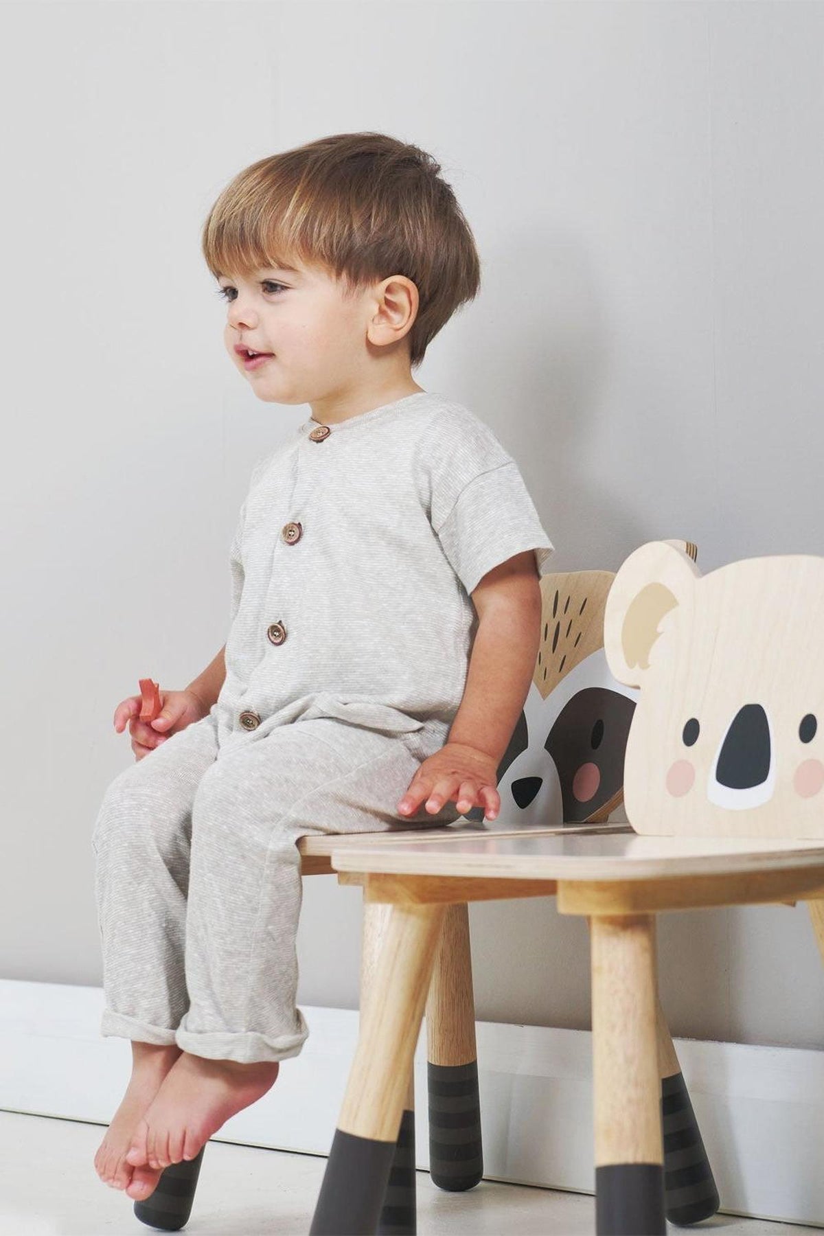 A toddler sits on a wooden chair featuring playful koala designs, wearing a light gray outfit with buttons.