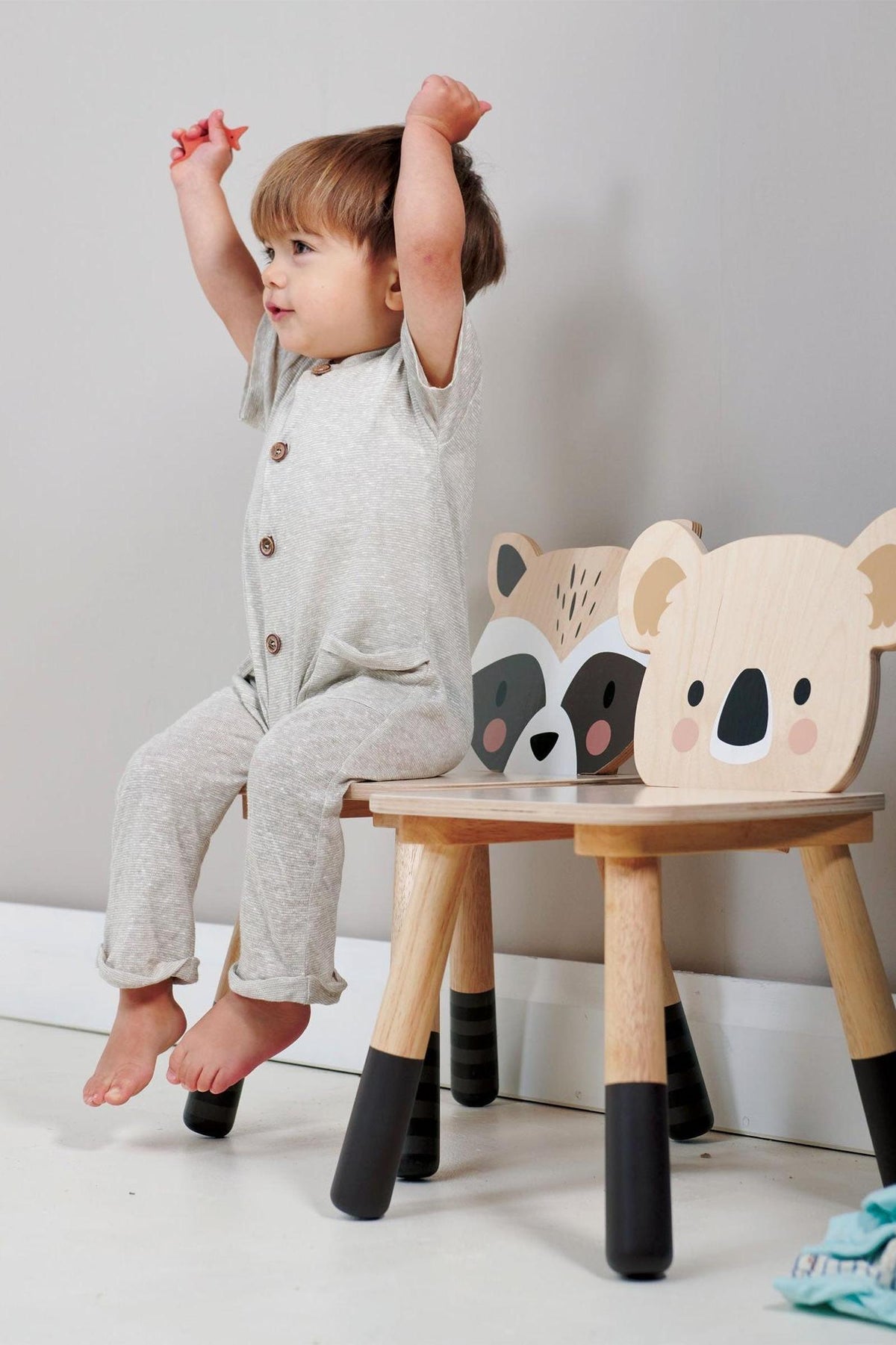 A toddler in a gray outfit sits on a wooden chair with raccoon and koala designs, smiling and raising arms.
