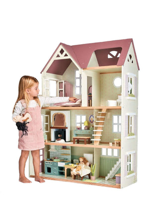 A young girl plays with a large, multi-story wooden dollhouse featuring detailed furniture and pastel colors.
