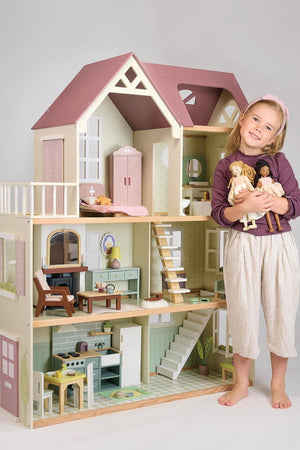 A young girl stands beside a large, multi-level wooden dollhouse, holding two dolls, smiling.