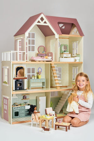 A young girl plays with a large, multilevel wooden dollhouse featuring pastel colors and miniature furniture.