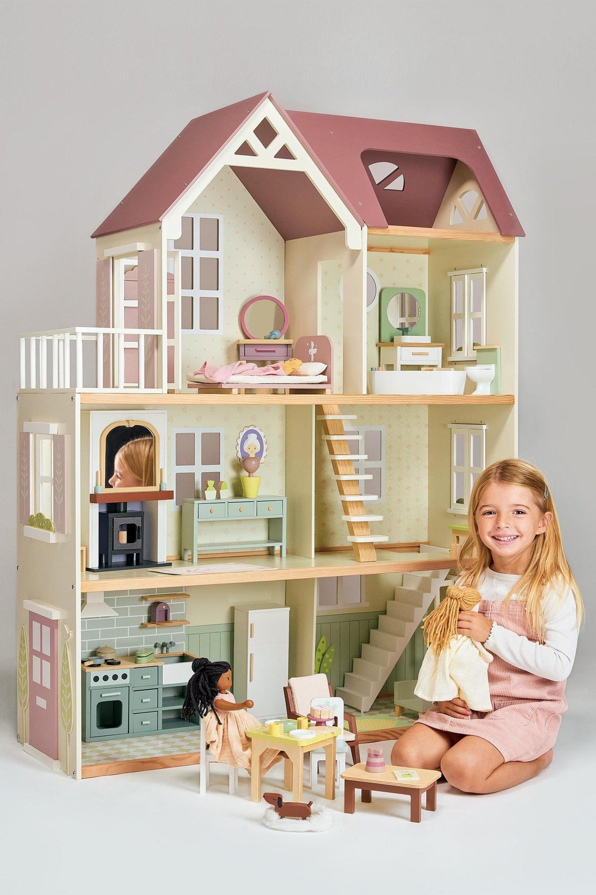 A young girl sits in front of a colorful wooden dollhouse, holding a doll outfit. The dollhouse features multiple rooms with detailed furnishings, showcasing a playful and inviting design.