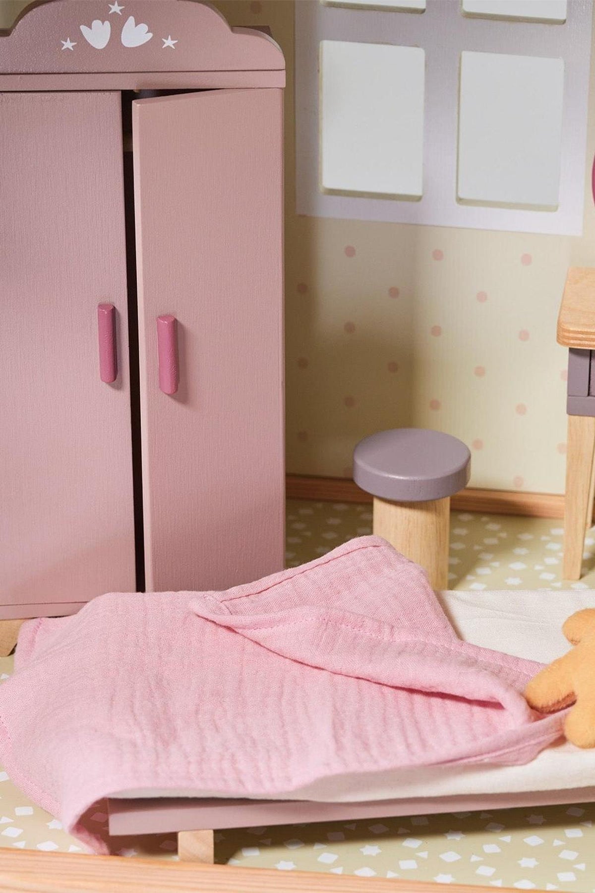 Wooden dollhouse interior with a pink wardrobe, matching blanket, and a small stool, set on a patterned floor.