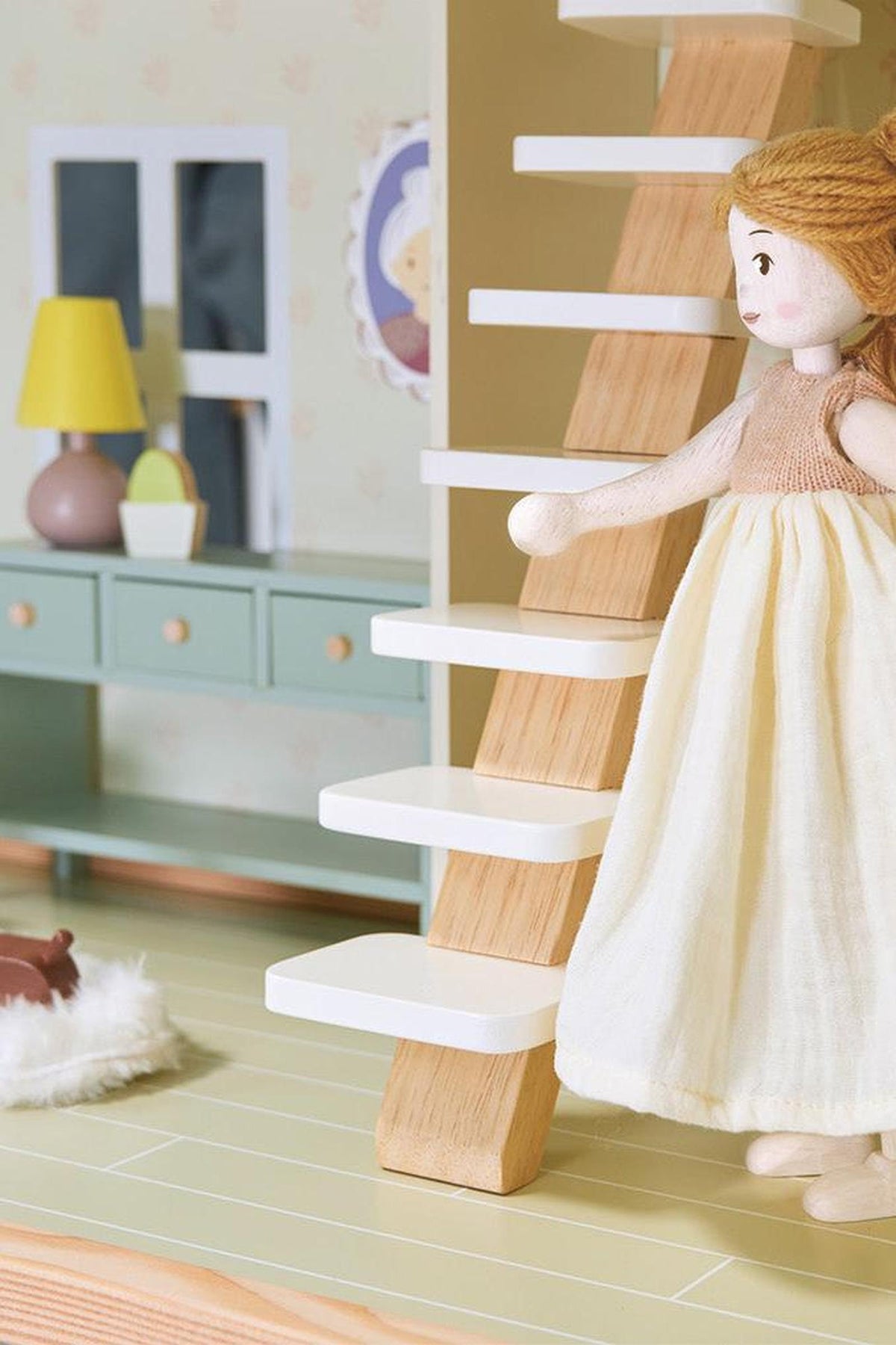 A wooden doll with a cream dress stands by a miniature staircase in a dollhouse setting.
