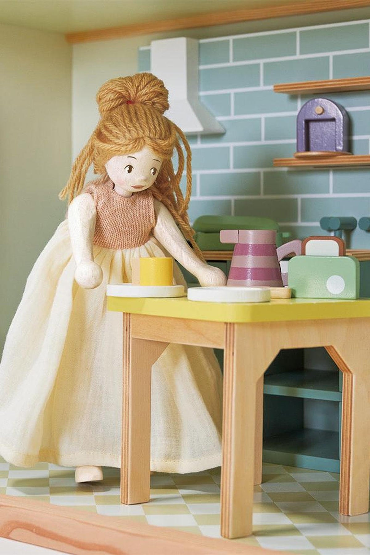 A handcrafted wooden doll with long hair, wearing a light dress, stands at a colorful kitchen table, interacting with vibrant play kitchen accessories, showcasing sustainable materials and imaginative play.