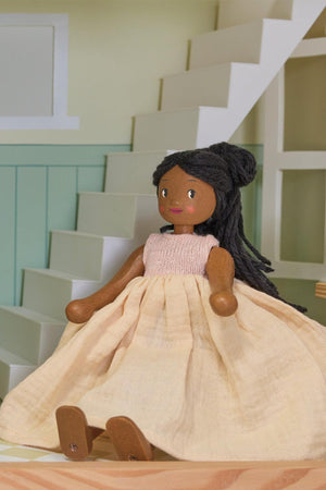 Wooden doll with dark hair and beige dress sits on stairs in a dollhouse setting.