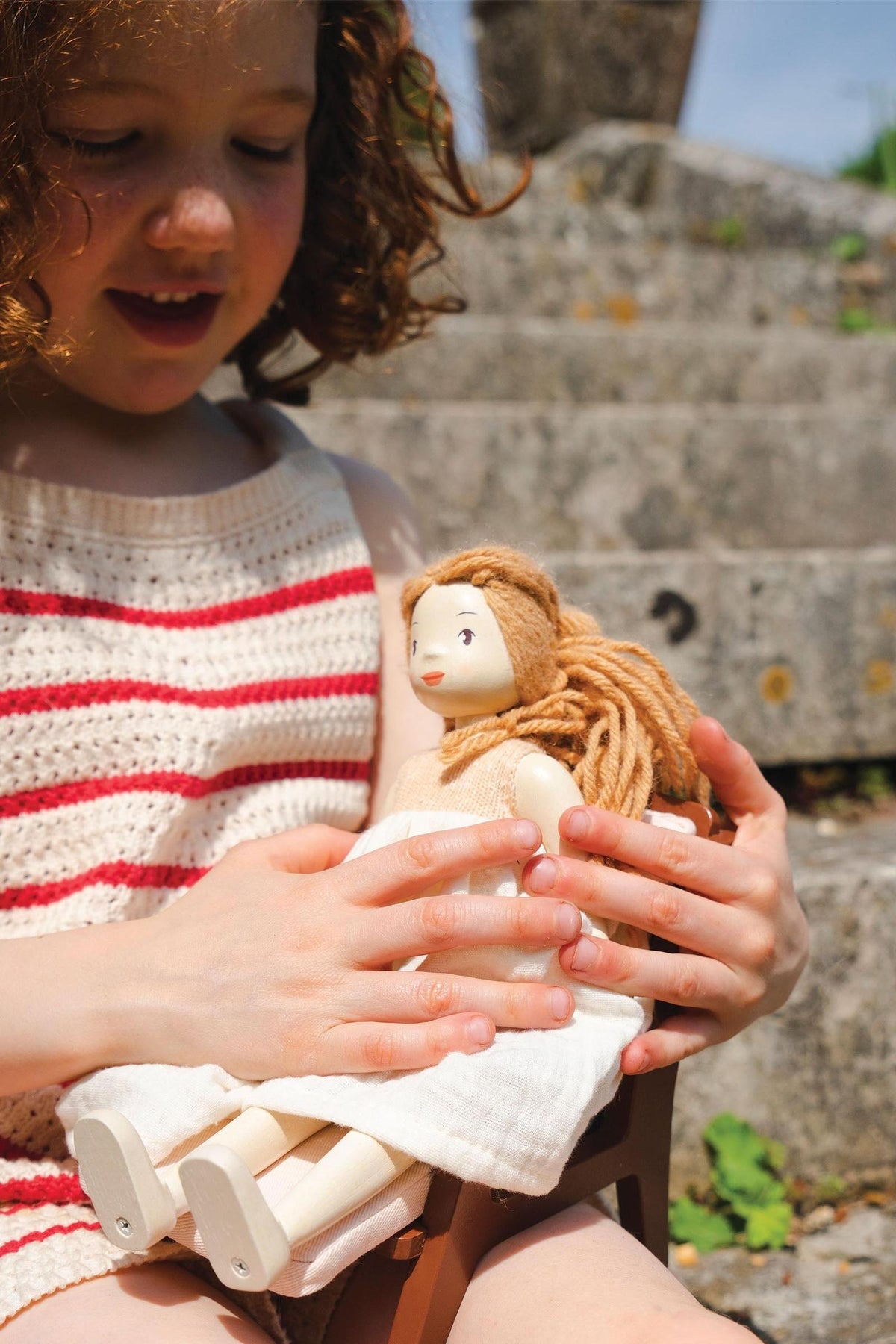 A young girl with curly hair sits outdoors, gently holding a handcrafted wooden doll with yarn hair. The doll features a natural wood finish and a simple, charming design.
