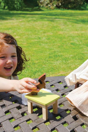 A child with curly hair plays outdoors, interacting with a colorful wooden toy on a small table. The setting features lush green grass, creating a cheerful and playful atmosphere.