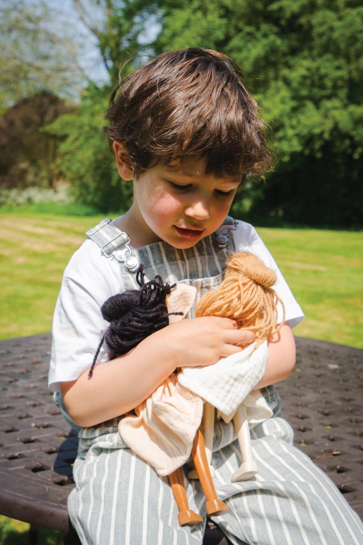 A young child sits outdoors, gently holding three handcrafted wooden dolls. The child wears a striped outfit and appears focused, surrounded by lush greenery in a sunny setting.