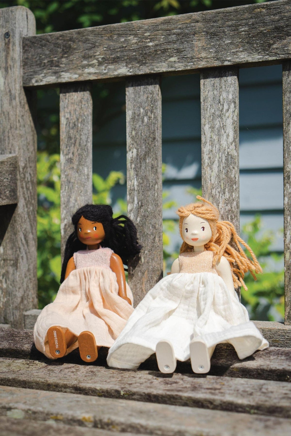 Two handcrafted wooden dolls sit on a rustic wooden bench. One doll has dark hair and a peach dress, while the other features blonde hair and a cream-colored dress.