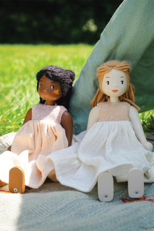 Two handcrafted wooden dolls sit on a blanket in a grassy setting. One doll has dark skin and black hair, while the other features light skin and blonde hair, both.