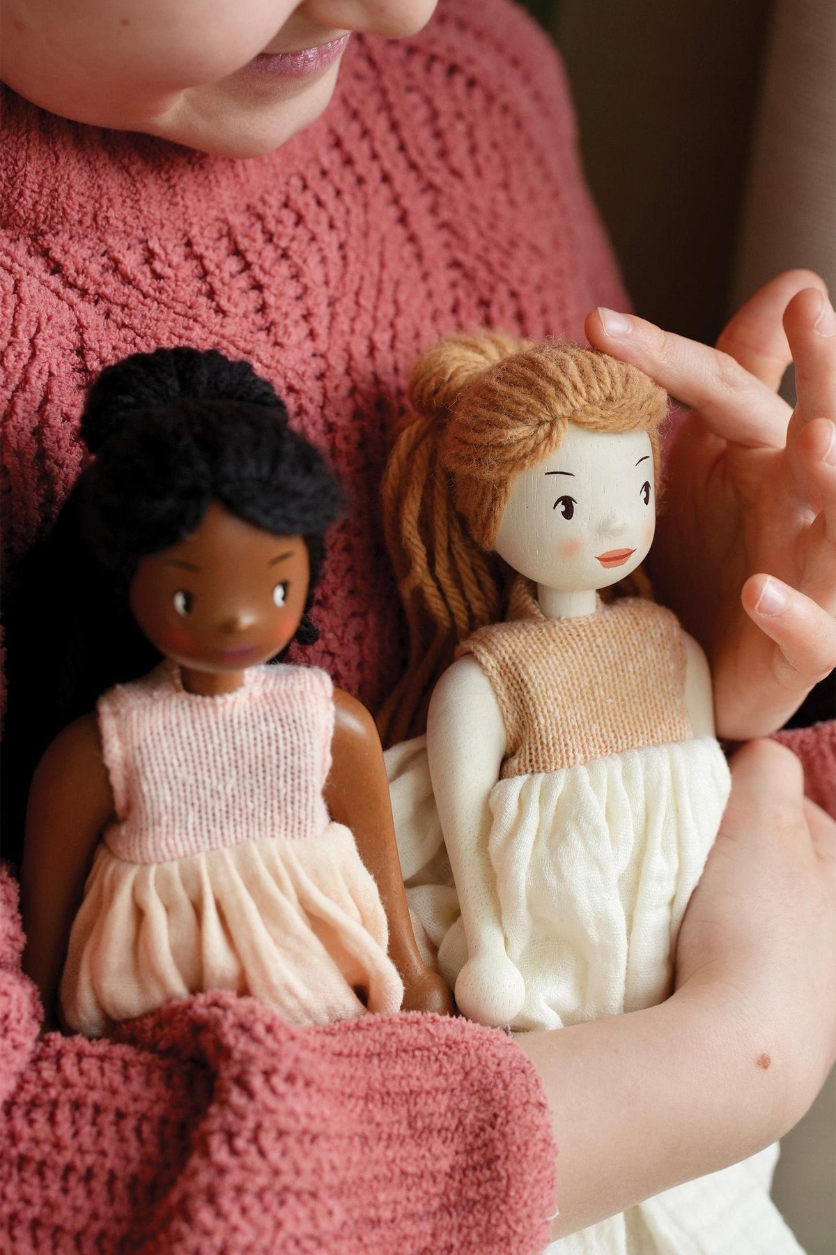 A child holds two handcrafted dolls with diverse features, wearing soft, pastel dresses. The dolls are made from sustainable materials, highlighting their eco-friendly design and charm.