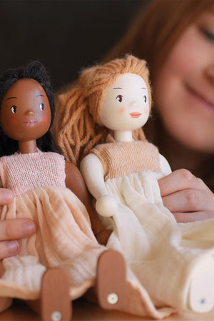 Two handcrafted wooden dolls with diverse skin tones are held by a child, showcasing natural yarn hair and soft, organic cotton dresses in muted colors.