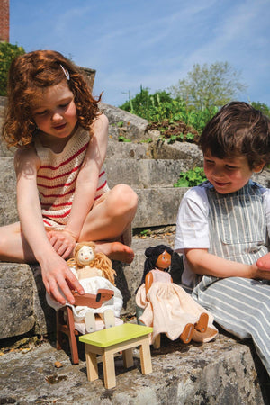 Two children sit on stone steps, engaging with wooden dolls. The scene features a light-colored doll with blonde hair and a dark-haired doll, alongside a small green table.