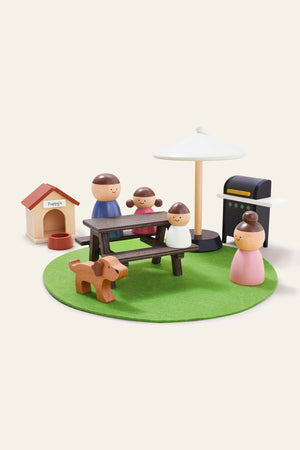 A colorful wooden playset featuring four family figures, a picnic table, a dog, a grill, and a playhouse, all set on a green felt mat, encouraging imaginative outdoor scenarios.