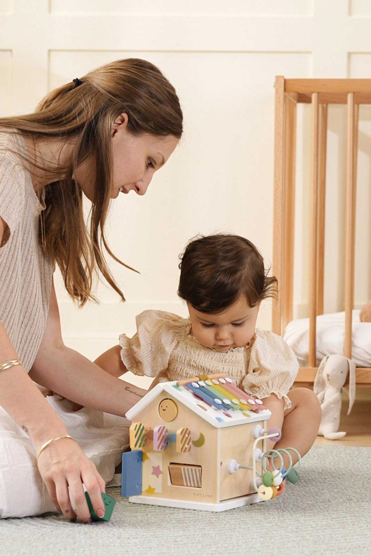 A mother and her toddler engage with a colorful wooden activity cube resembling a house, featuring musical elements and interactive parts, in a cozy, well-lit nursery setting.