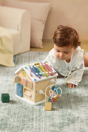 A toddler explores a colorful wooden activity cube with various interactive features, set on a soft green rug.