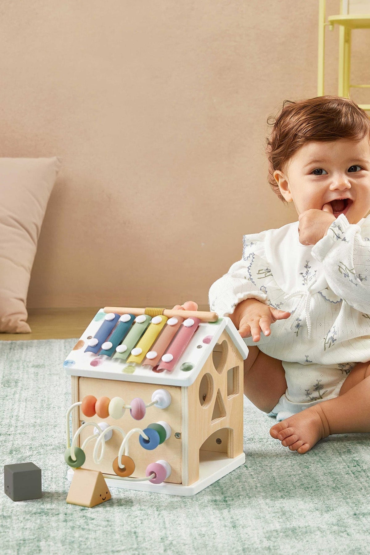 A smiling toddler sits on a soft green rug, interacting with a colorful wooden activity cube featuring a xylophone, shape sorter, and beads, promoting hands-on learning and motor skills.