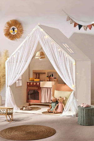 A cozy playhouse features sheer white curtains, a wooden kitchen, and plush toys, creating a warm, inviting space for children.