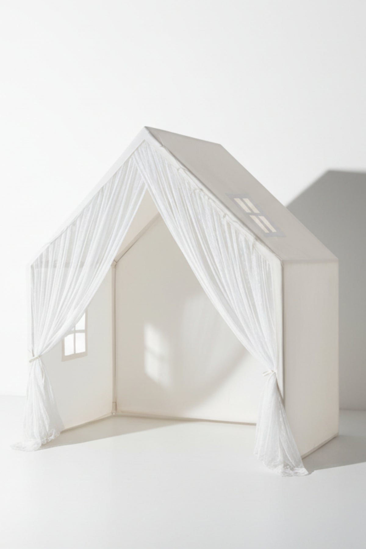 A white playhouse with sheer curtains and window cutouts, designed for children’s imaginative play in a bright, minimal setting.