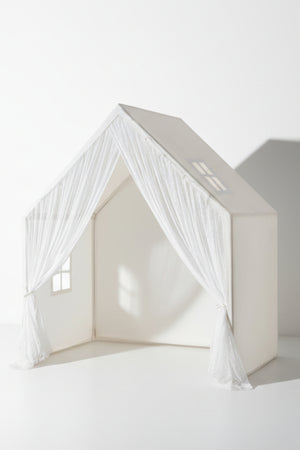 A white playhouse with sheer curtains and window cutouts, designed for children’s imaginative play in a bright, minimal setting.