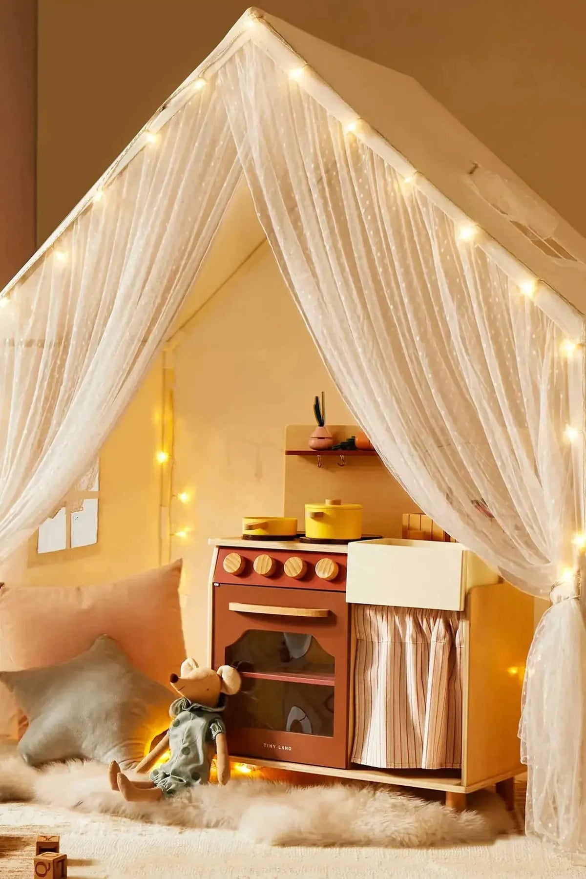 A cozy playhouse interior features soft curtains, a wooden kitchen set with yellow pots, and plush pillows on a fluffy.