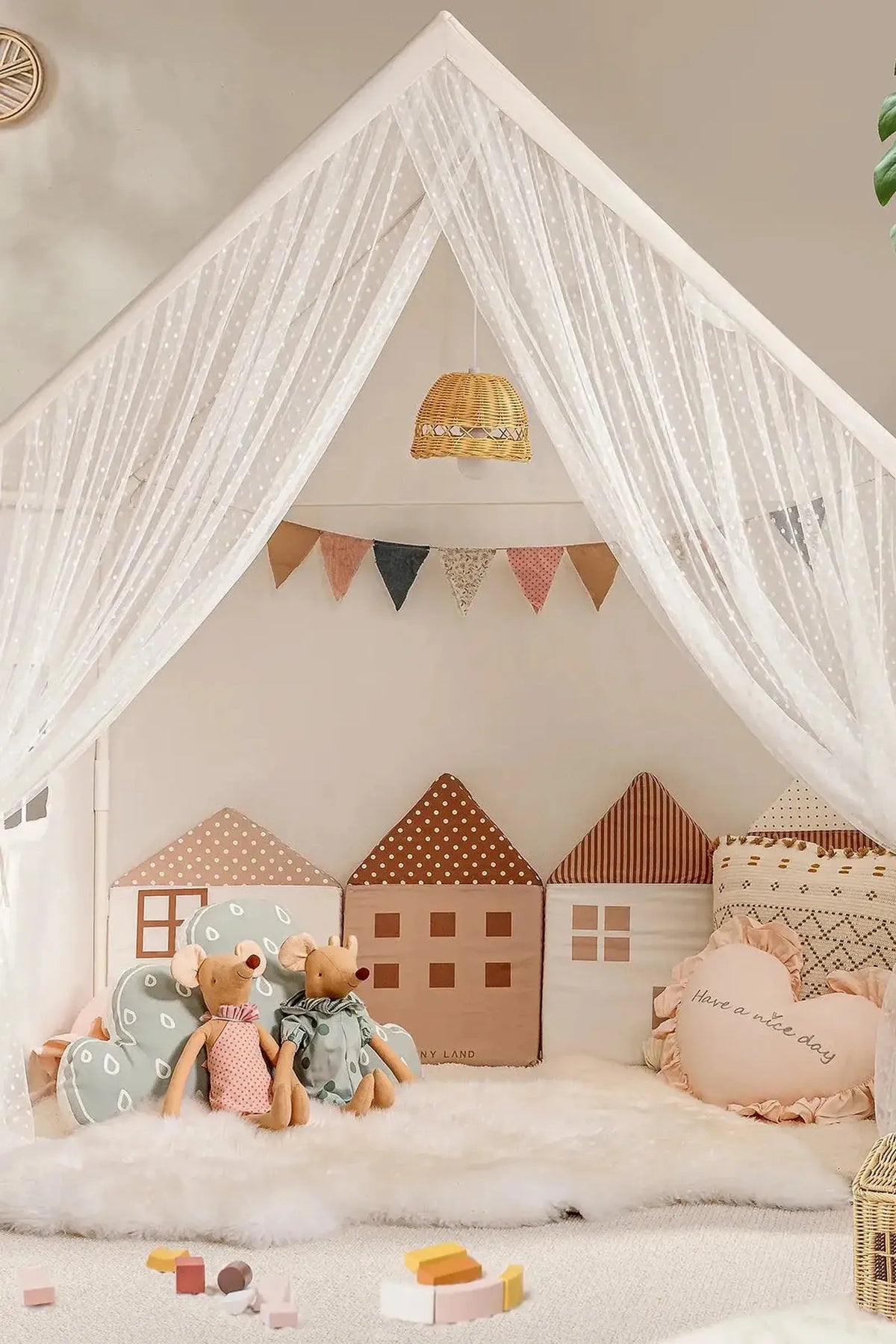 A cozy play area features soft plush cushions shaped like houses, two stuffed mice, and decorative bunting under sheer white.