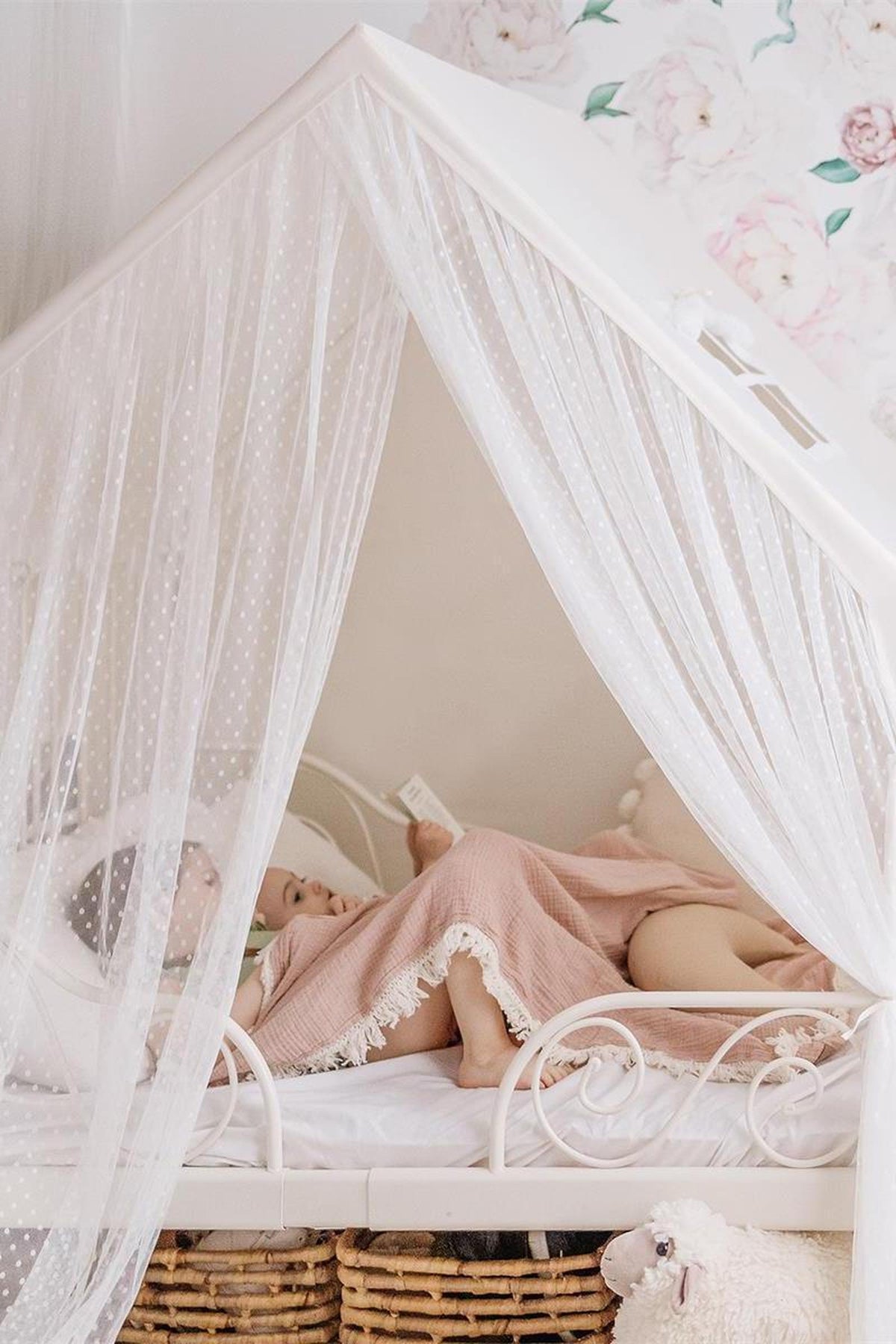 Two children relax under a sheer white canopy in a cozy bed, wrapped in a soft pink blanket.