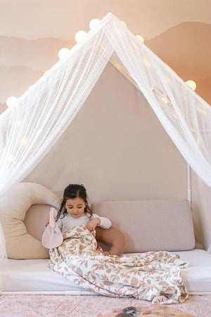 A young girl sits on a cozy sofa, wrapped in a floral blanket, surrounded by soft pillows and fairy lights.