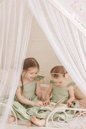 Two young girls in matching green dresses sit on a bed, reading a book under a sheer, polka-dotted canopy.