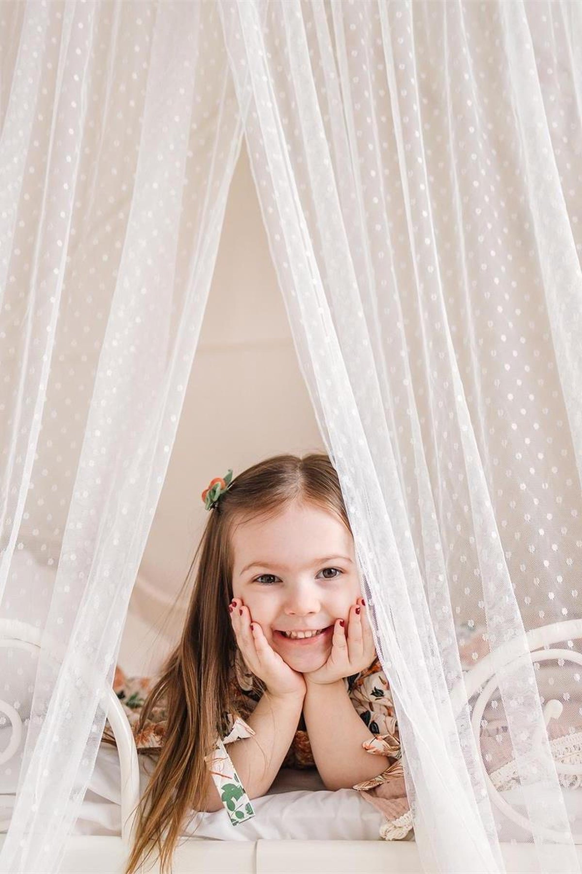 A smiling girl peeks through sheer, dotted white curtains while resting on a bed, showcasing her playful expression.
