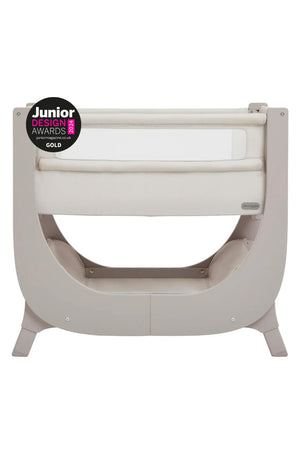 A Shnuggle Air Lite Bedside Bassinet features a sleek, curved design in soft taupe with a breathable mesh side. It is height-adjustable, designed for infant safety and comfort.