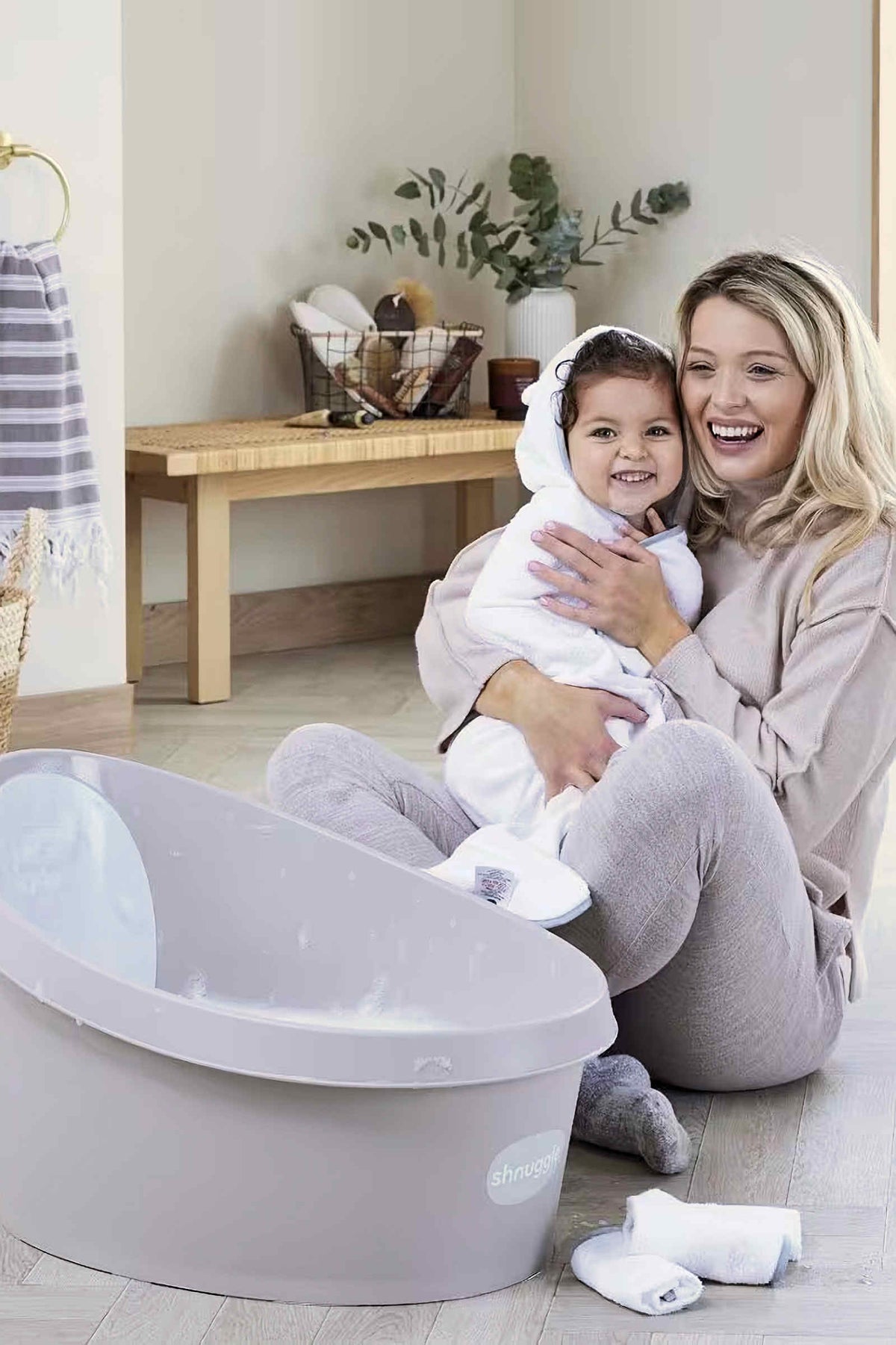 A mother and child smile beside a grey Shnuggle Toddler Bath Tub in a cozy bathroom setting.