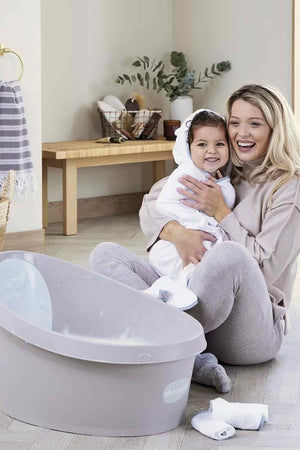 A mother and child smile beside a grey Shnuggle Toddler Bath Tub in a cozy bathroom setting.
