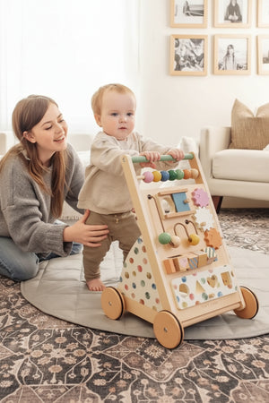 A toddler stands with support from a parent, using a colorful wooden baby walker adorned with various shapes and textures. The scene is set in a cozy, well-lit living room.