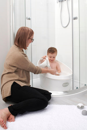 A child sits in a white Shnuggle Toddler Bath while an adult assists, inside a modern shower enclosure.