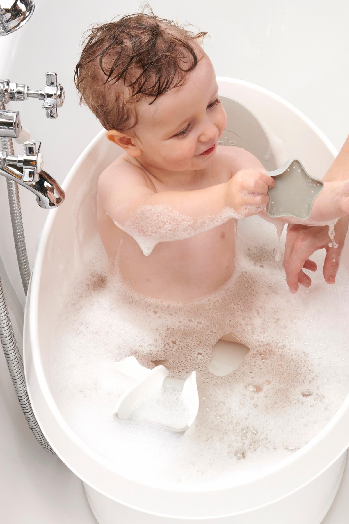 A toddler plays with a star-shaped toy in a white Shnuggle bath tub filled with bubbles.