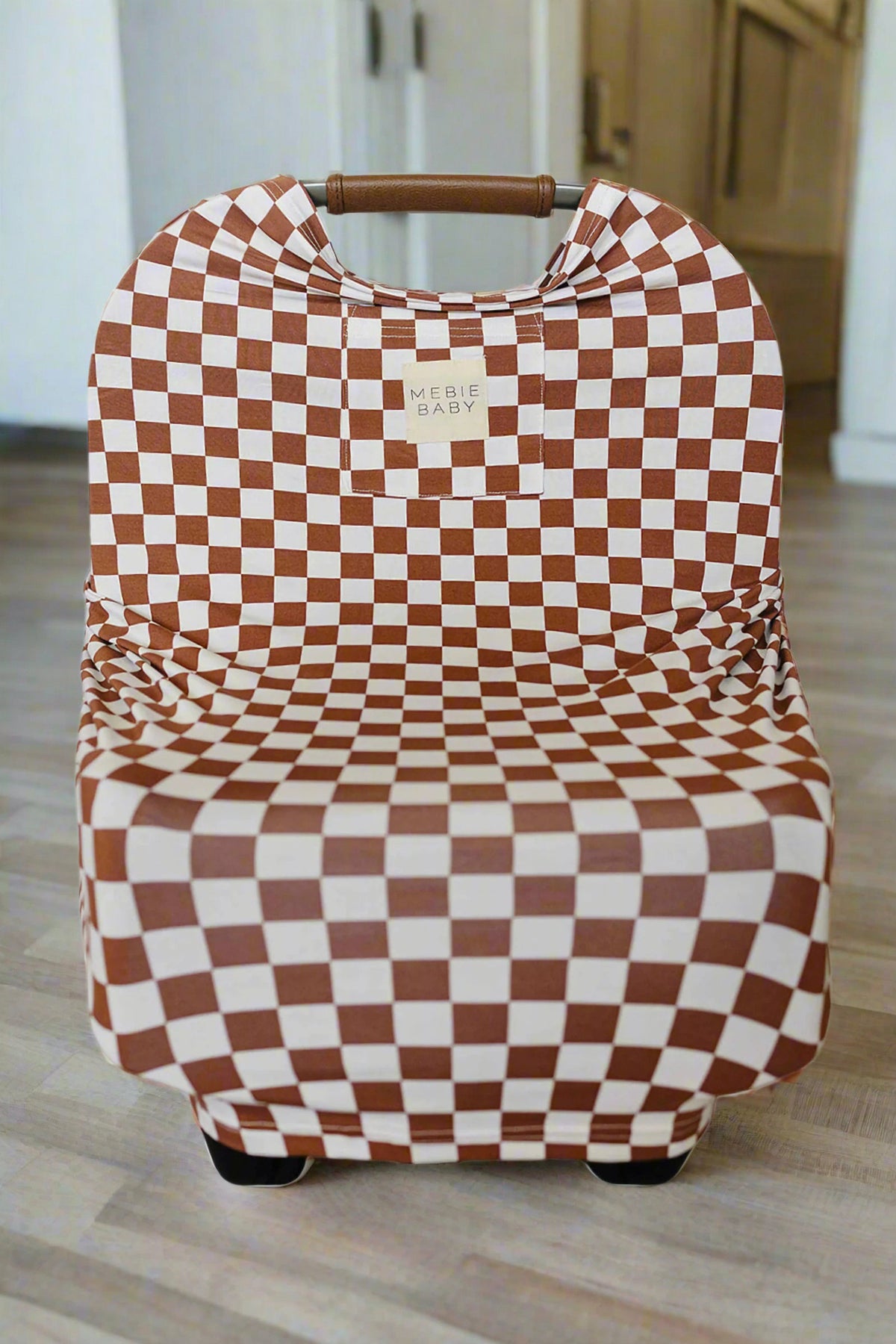 Rust checkered baby cover on car seat, featuring Mebie Baby label, in a light-colored room.